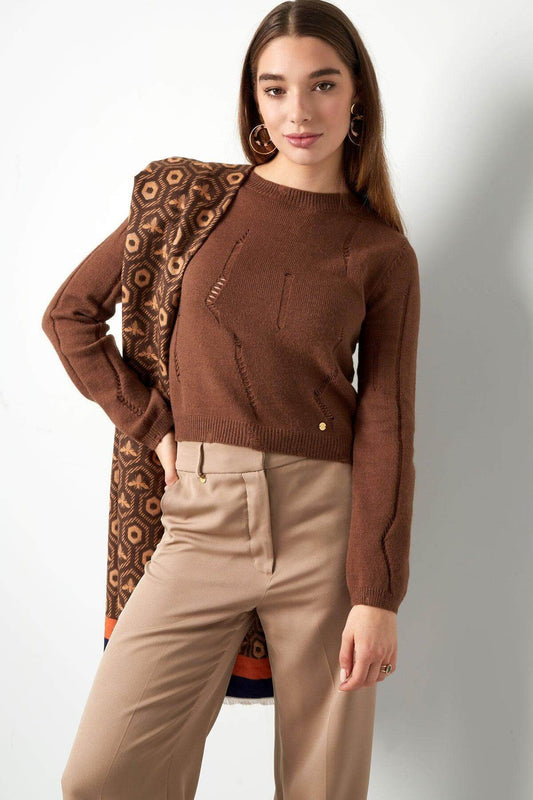 Woman wearing a brown knit pullover, paired with beige pants and holding a patterned scarf, showcasing cozy and stylish winter fashion.