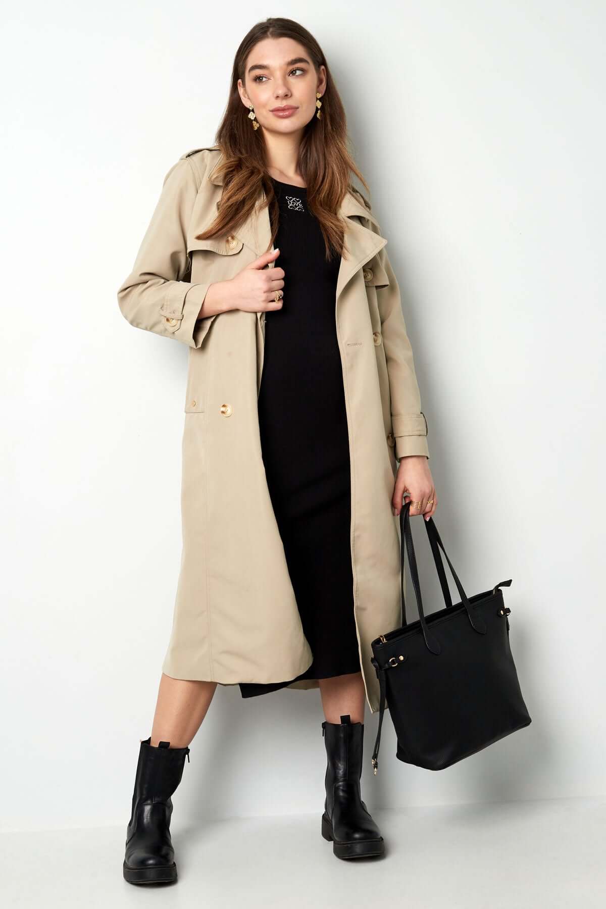 Woman in a beige trench coat over a black dress, holding a black handbag, stylish urban outfit.