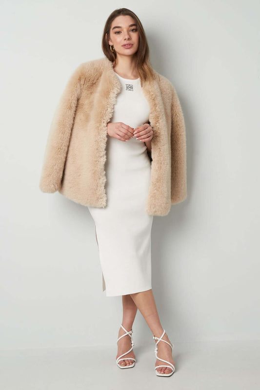 Elegant woman in a white fitted dress with a beige fur coat, showcasing sophisticated winter fashion.