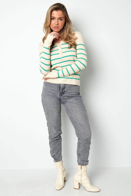 Woman in white and green striped V-neck sweater, paired with gray jeans and white boots, posing against white background.