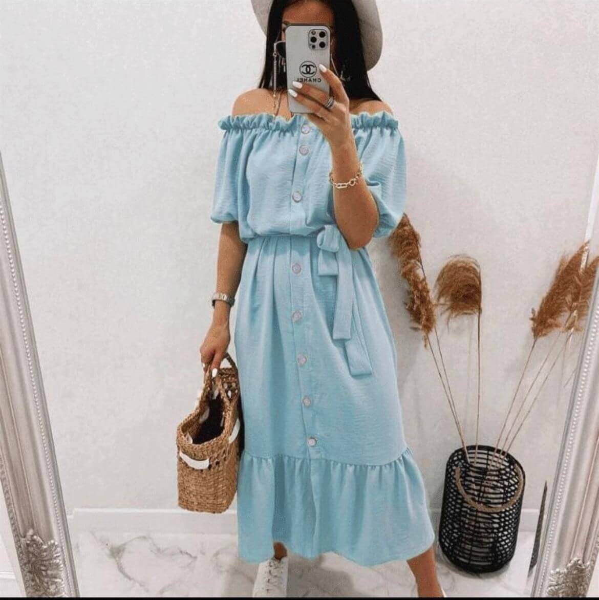 Woman in a light blue off-shoulder dress with buttons, standing in a mirror, holding a straw bag.